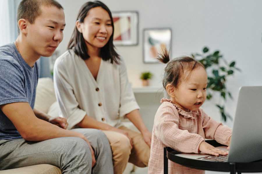 Parents smiling as their toddler taps at a laptop on a low table